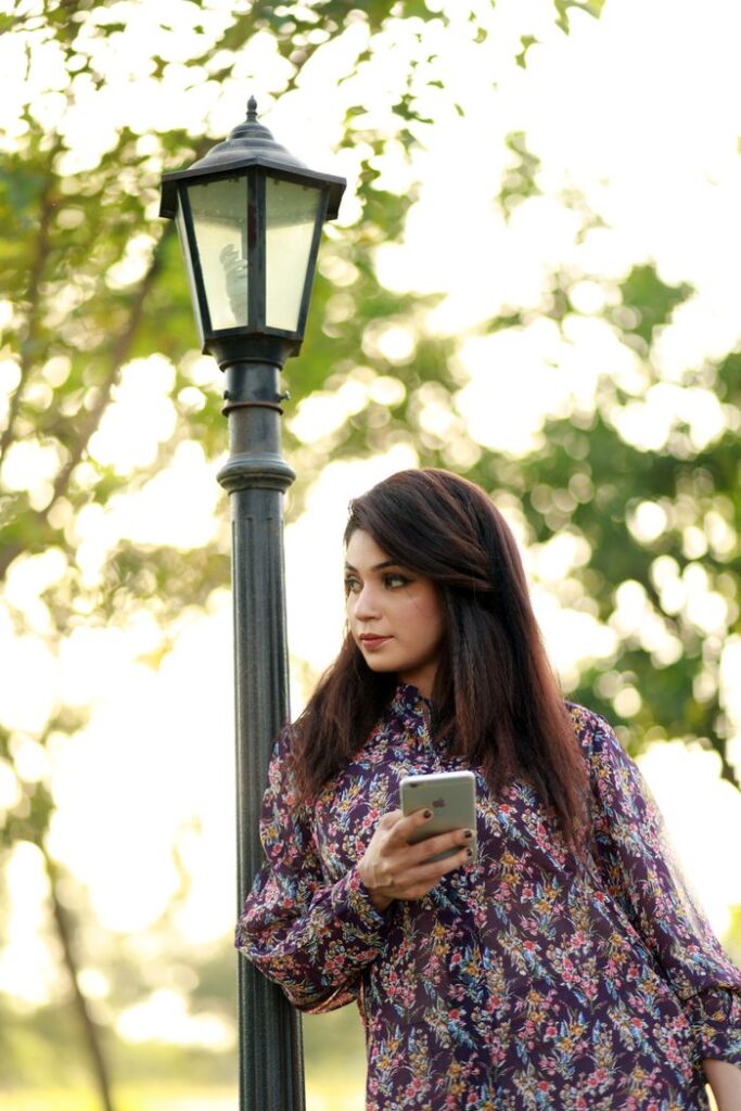 Stylish woman in a floral dress holding a smartphone by a lamp post outdoors.