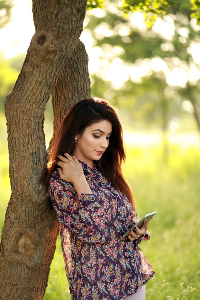 Chic woman leaning against a tree trunk using a smartphone outdoors on a sunny day.