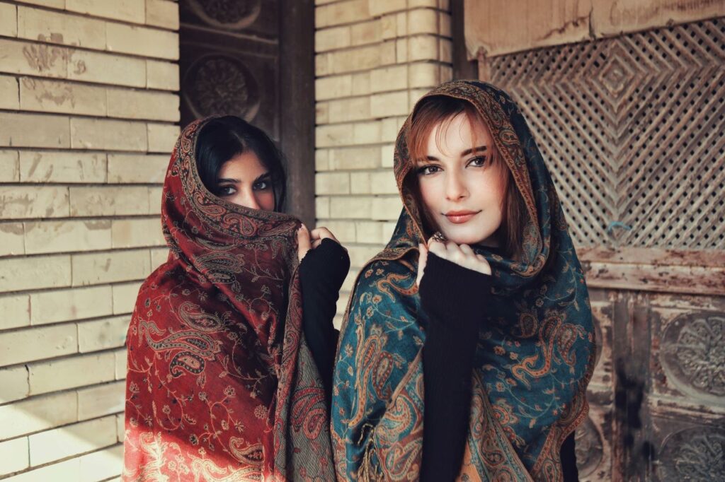 Two women wrapped in vibrant patterned scarves pose indoors with a vintage backdrop.