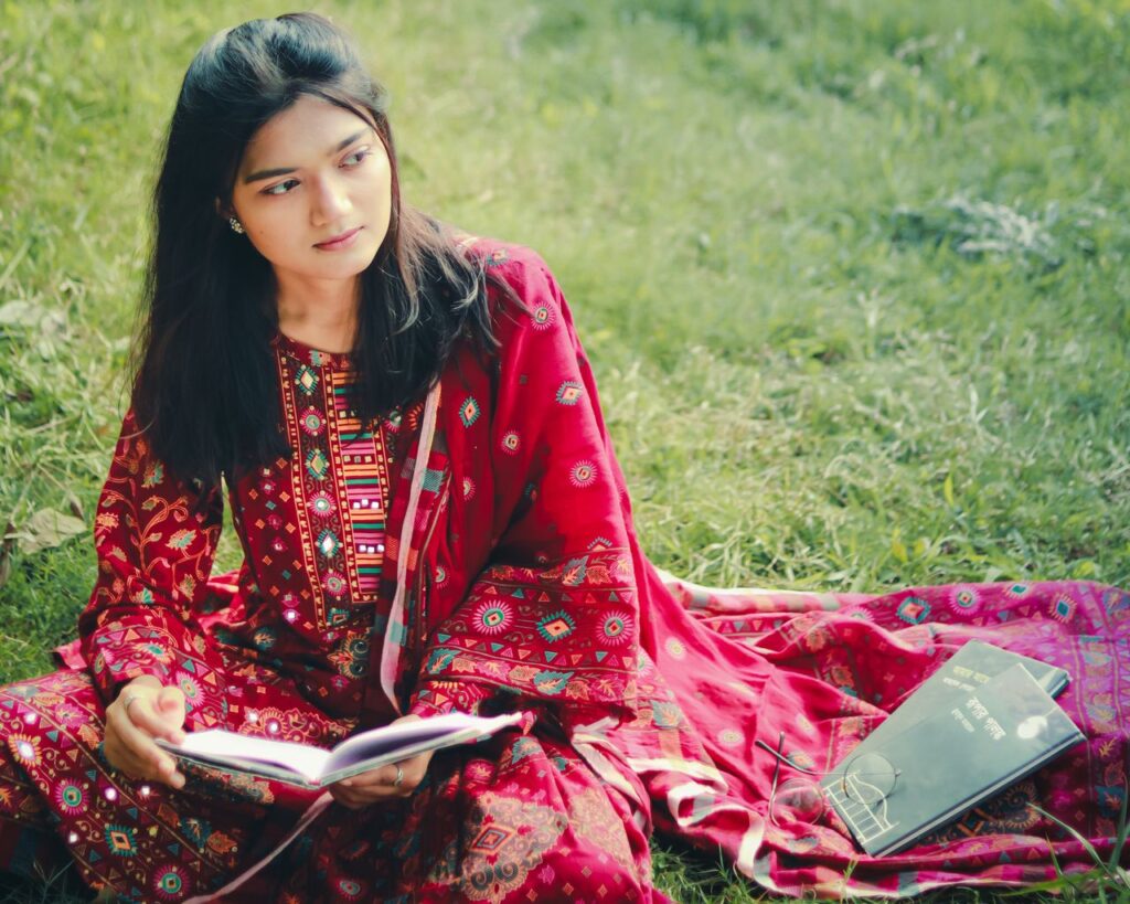 A young woman in a vibrant traditional outfit reading a book while sitting on grass in Khulna, Bangladesh.